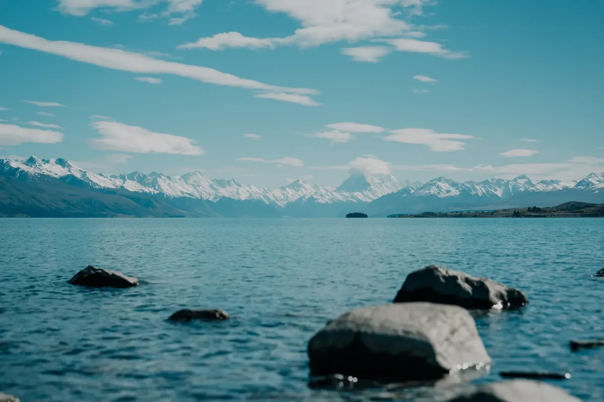 Tranquil lake with snowy mountains in New Zealand under a bright sky.