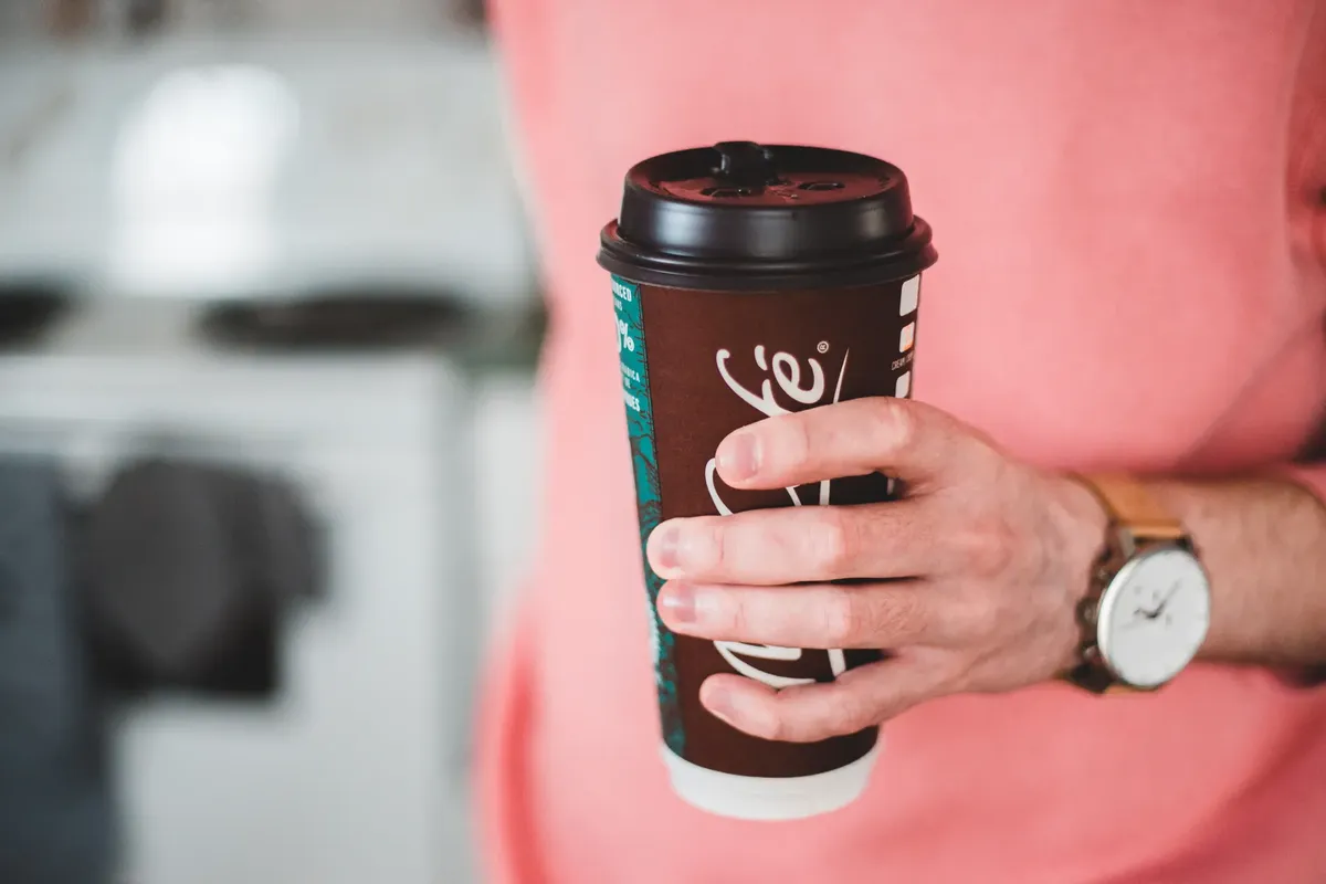 A person wearing a watch holds a takeaway coffee cup indoors with a blurred background.