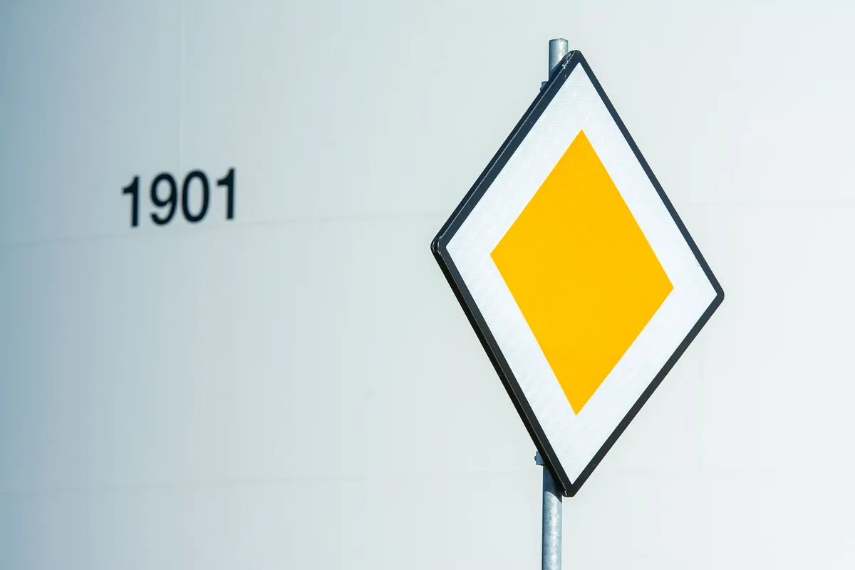 Close-up of a diamond-shaped yellow priority road sign against an industrial backdrop with the number 1901 visible.